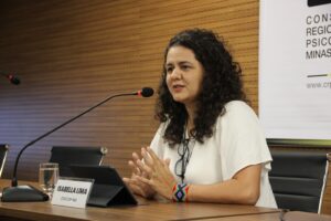 Foto da conselheira Isabella Lima sentada à mesa do auditório, falando ao microfone