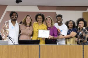 Foto mostra Dalcira recebendo a homenagem na ALMG. Ela está de pé, rodeada por deputadas e outras pessoas presentes ao evento.